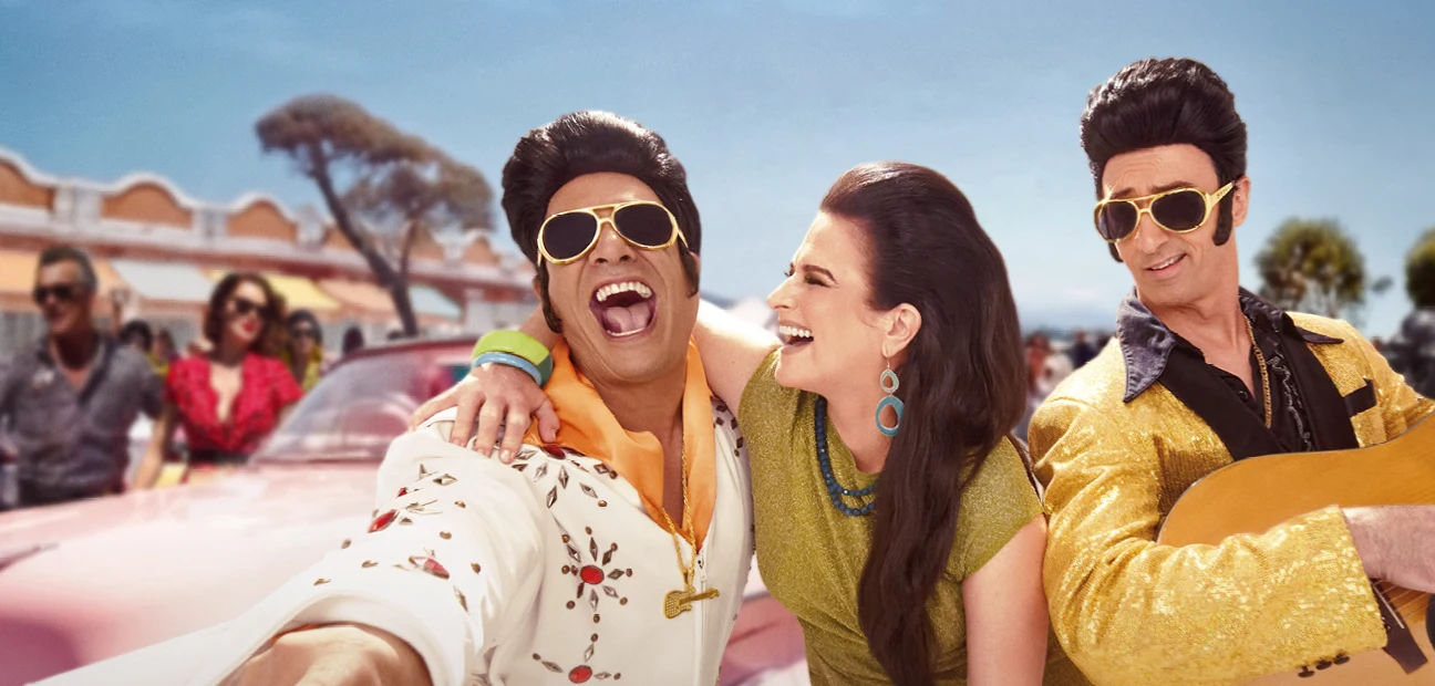 Two Elvis performers and a woman laughing together in front of a pink Cadillac, with a crowd and a showground building behind them