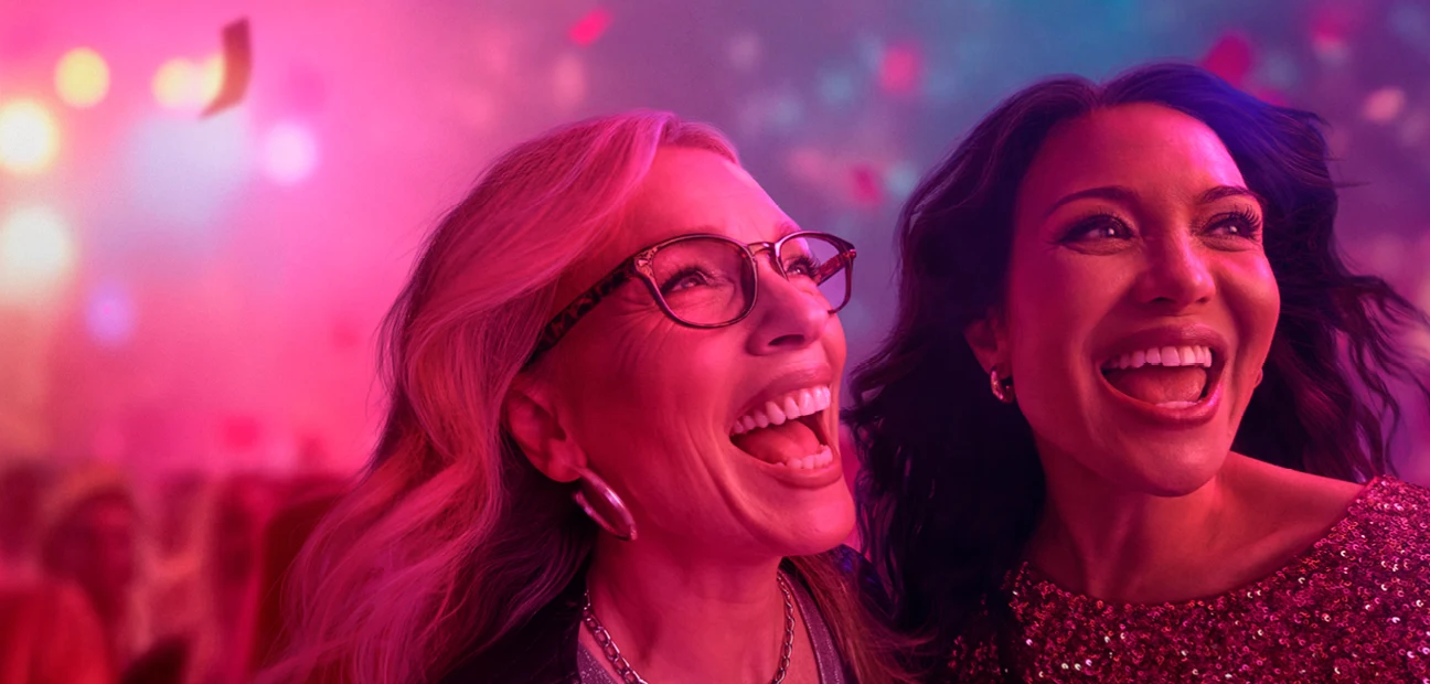 Worth Doing Well visual: two women laughing together in the crowd at a live music event under pink stage light