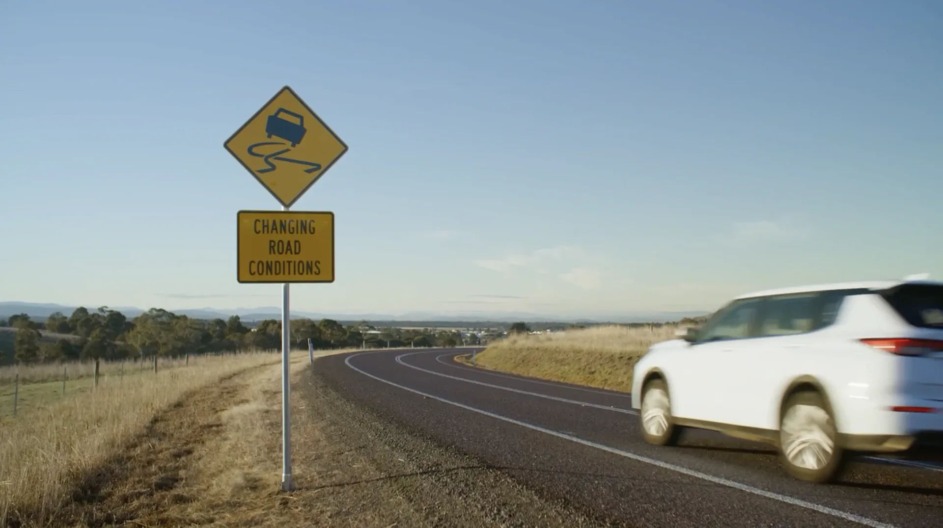 A yellow Changing Road Conditions sign beside a rural Tasmanian highway as a white car passes in daylight