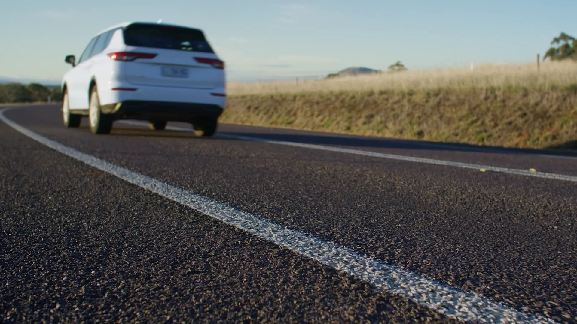 A white SUV on a rural Tasmanian highway in low afternoon sun, shot from road level with the line marking in the foreground